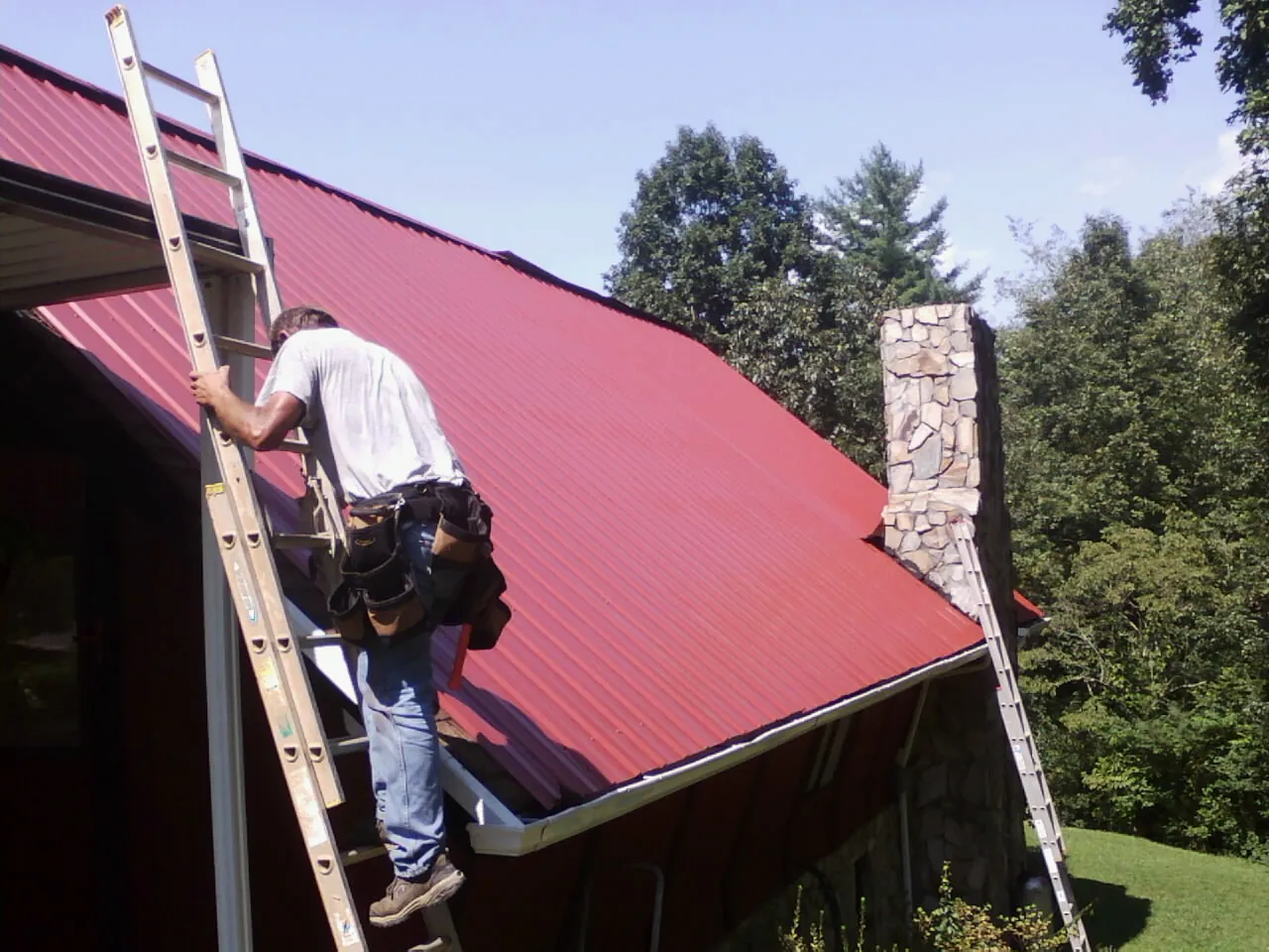 Roofer on a ladder beside a red metal roof and stone chimney — Soffit & Fascia Repair by Roofing Brothers in Highlands